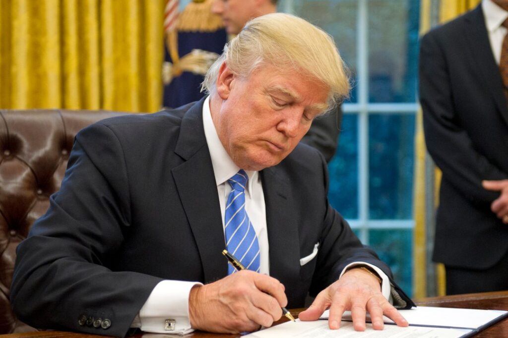 Person in a suit signing a document at a desk in an office setting.