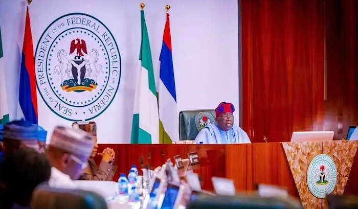 Meeting room with flags and emblem of Nigeria; a person sits at a desk with "President" on a plaque, surrounded by others at tables.