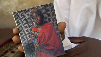 A hand holding a photo of a woman in a red headscarf and traditional attire, set against a geometric patterned background.