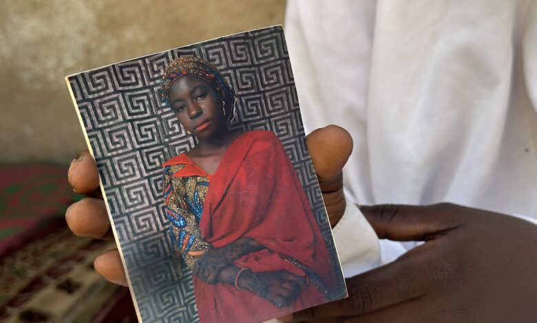 A hand holding a photo of a woman in a red headscarf and traditional attire, set against a geometric patterned background.