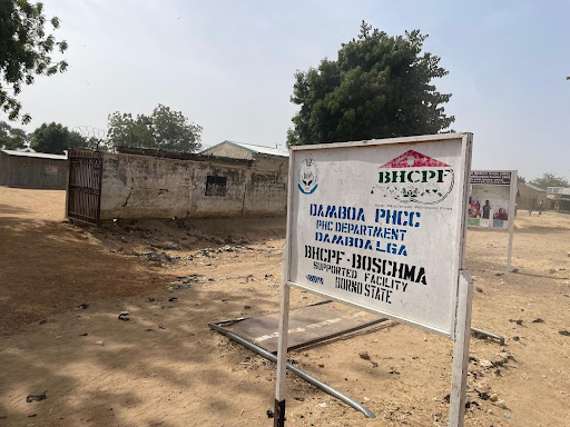 Sign for Damboa PHCC in Dacabra, Borno State, stands in a dusty area with trees and a building in the background.