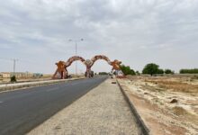 Wide road with a large, rusty archway and barren landscape; cloudy sky overhead.