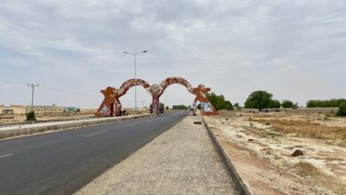 Wide road with a large, rusty archway and barren landscape; cloudy sky overhead.