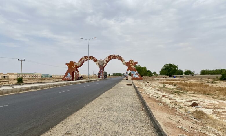 Wide road with a large, rusty archway and barren landscape; cloudy sky overhead.