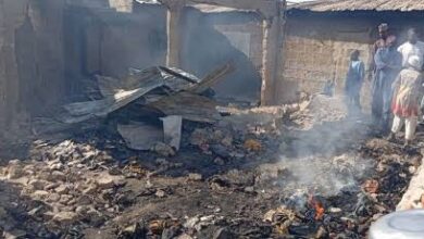 Burned debris and rubble in front of a damaged building, with several people observing the scene.