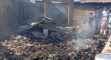 Burned debris and rubble in front of a damaged building, with several people observing the scene.