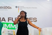 Person using crutches speaks into a microphone at a podium with NG Youth SDGs and German Embassy logos in the background.