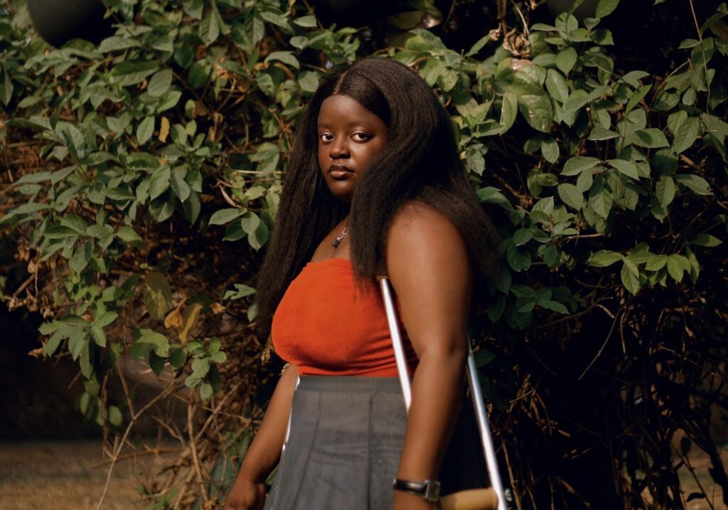 A woman with long hair, wearing an orange top and gray skirt, stands using crutches in front of lush green foliage.