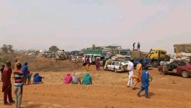 People and vehicles gather on a dusty road, some sitting, waiting, while others stand and walk around. Trucks and cars are parked closely.