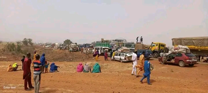 People and vehicles gather on a dusty road, some sitting, waiting, while others stand and walk around. Trucks and cars are parked closely.