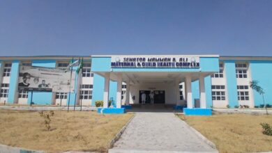 Blue and white building of the Senator Mamman B. Ali Maternal & Child Health Complex, with a billboard and flags in the foreground.