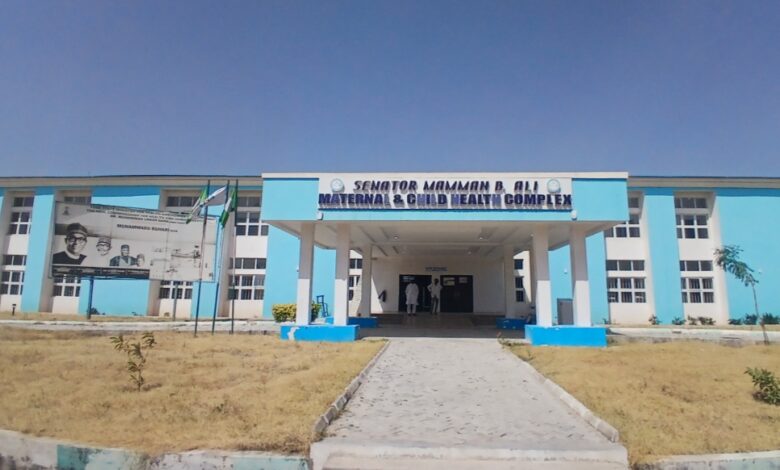 Blue and white building of the Senator Mamman B. Ali Maternal & Child Health Complex, with a billboard and flags in the foreground.