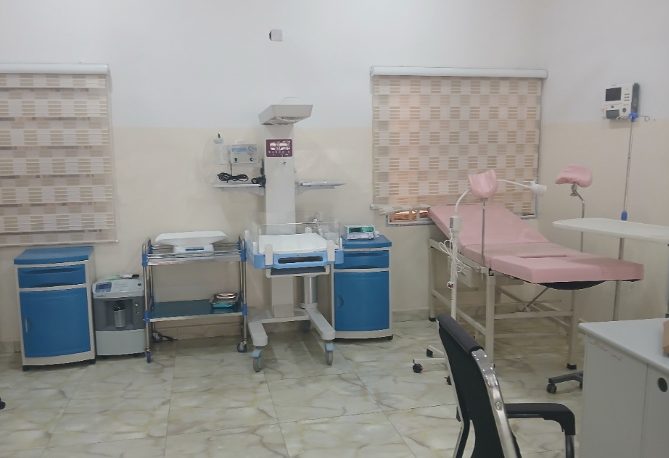 A medical room with examination bed, incubator, blue cabinets, and medical equipment against cream walls and tiled floor.