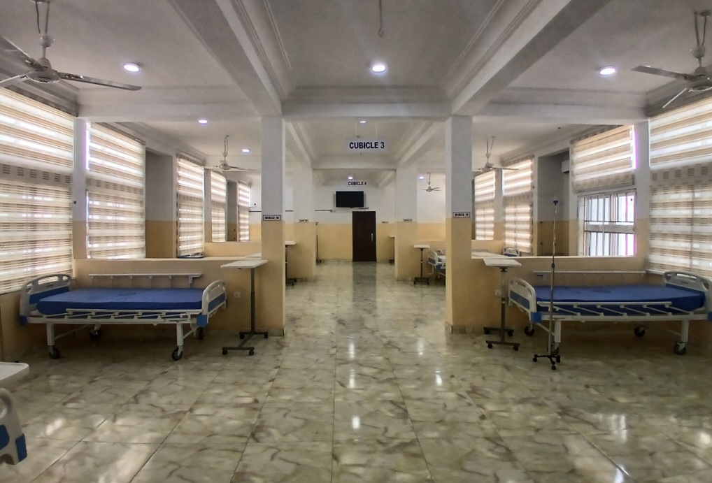 Empty hospital ward with multiple beds and IV stands, bright lighting, and tiled flooring.