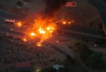 Aerial view of a large fire on a highway at night, with vehicles nearby and dark smoke rising into the sky.