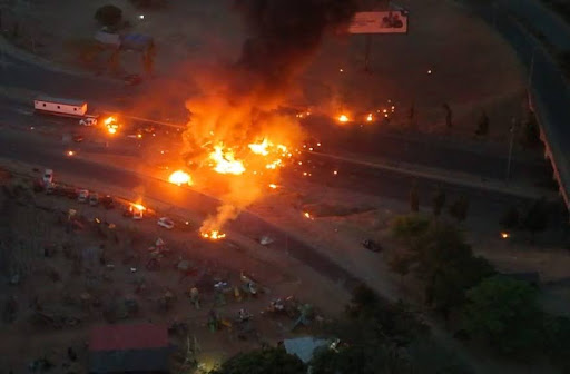 Aerial view of a large fire on a highway at night, with vehicles nearby and dark smoke rising into the sky.