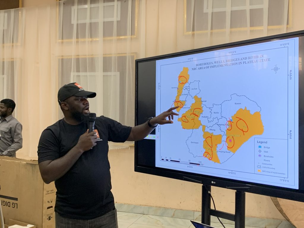 Man presenting a map highlighting boreholes, wells, and rivers in Plateau State on a screen, pointing with a microphone in hand.