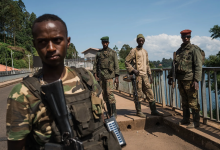 Four soldiers in camouflage stand on a bridge holding rifles, with trees and a river in the background.