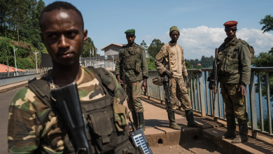 Four soldiers in camouflage stand on a bridge holding rifles, with trees and a river in the background.