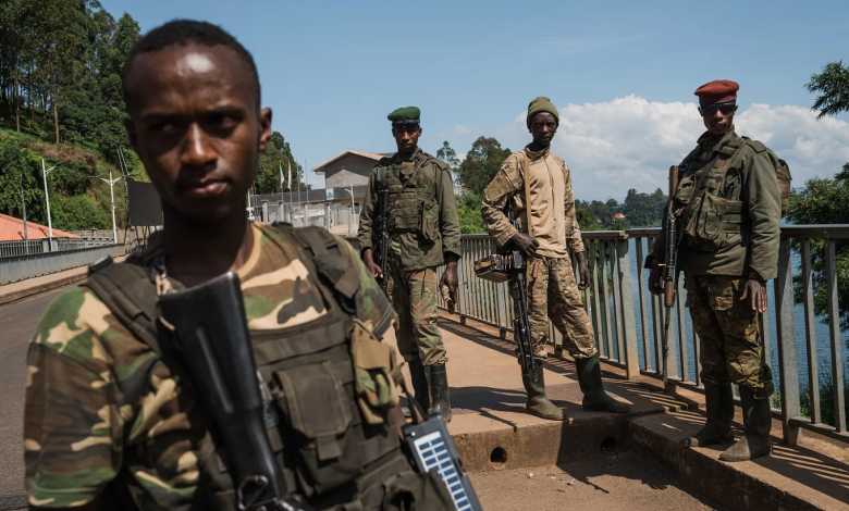 Four soldiers in camouflage stand on a bridge holding rifles, with trees and a river in the background.