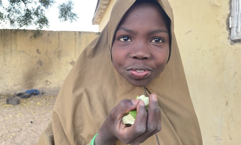 A person in a brown shawl holds a green fruit, standing outside next to a yellow wall.