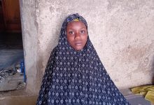 A person in a patterned dark blue hijab stands against a textured wall.