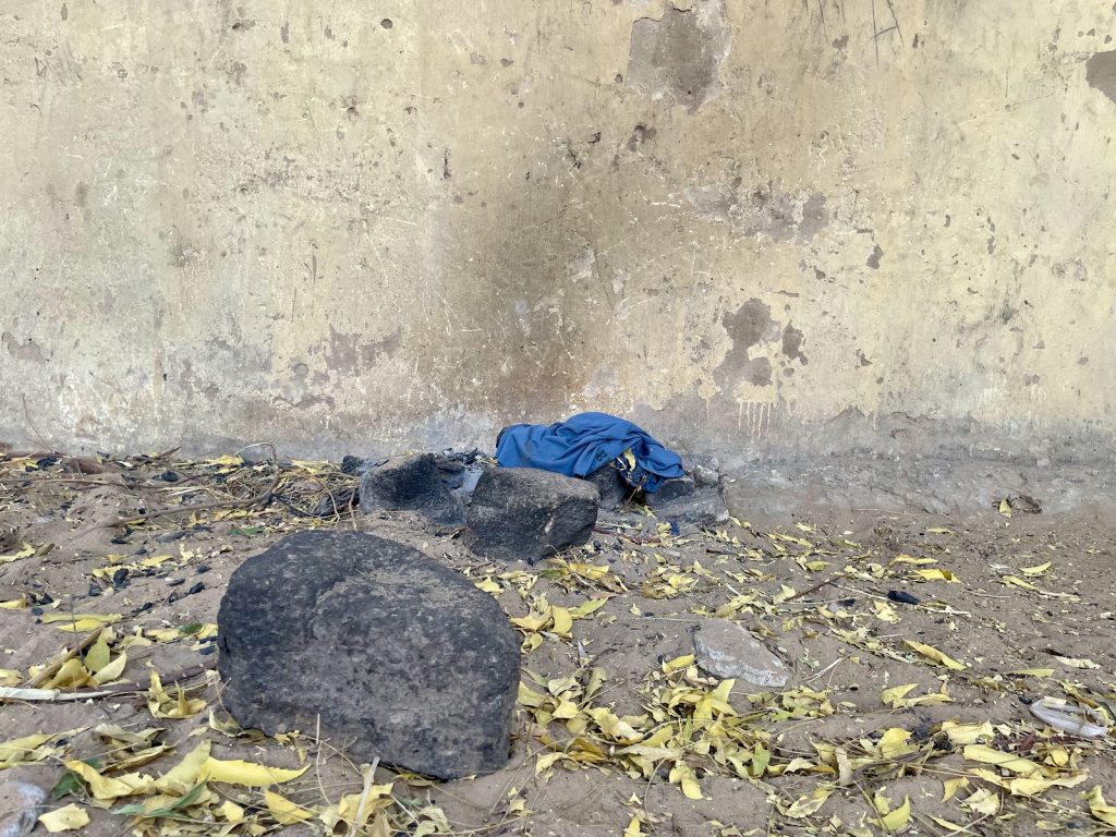 Ground with dry leaves and rocks in front of a worn wall. A blue shirt lies on the ground near the wall.