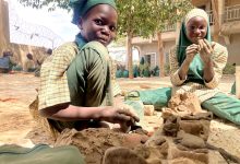 Children in uniforms engage in clay modeling outdoors, surrounded by trees and a school building.