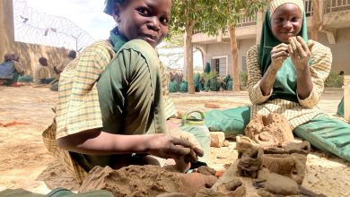 Children in uniforms engage in clay modeling outdoors, surrounded by trees and a school building.