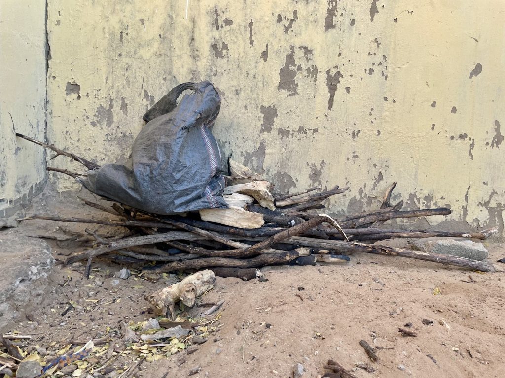 A pile of sticks and a black bag rest against a weathered, peeling wall on a dirt ground.