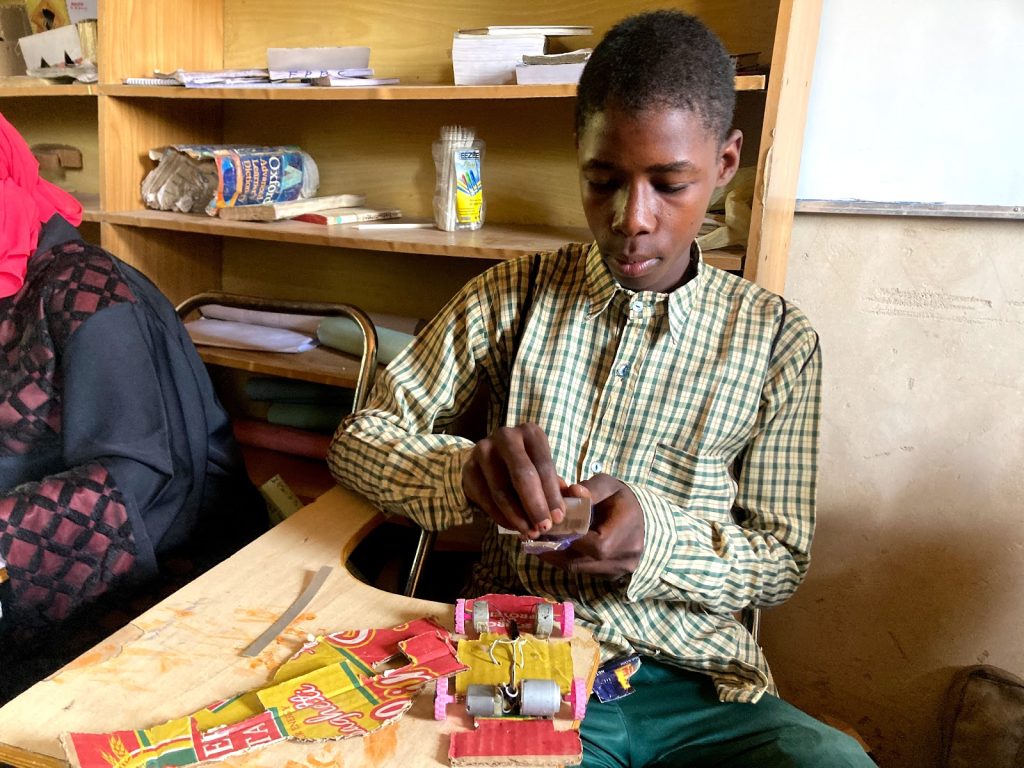 Boy in a checkered shirt concentrates on crafting with small materials at a desk, with books and supplies on shelves behind him.