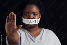 Person with tape labeled "violence" over mouth, holding hand out in a stop gesture against a dark background.