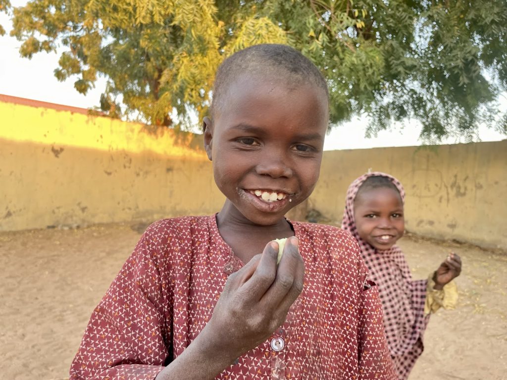 Two smiling children stand outdoors, one holding a small object. A tree and wall are in the background.