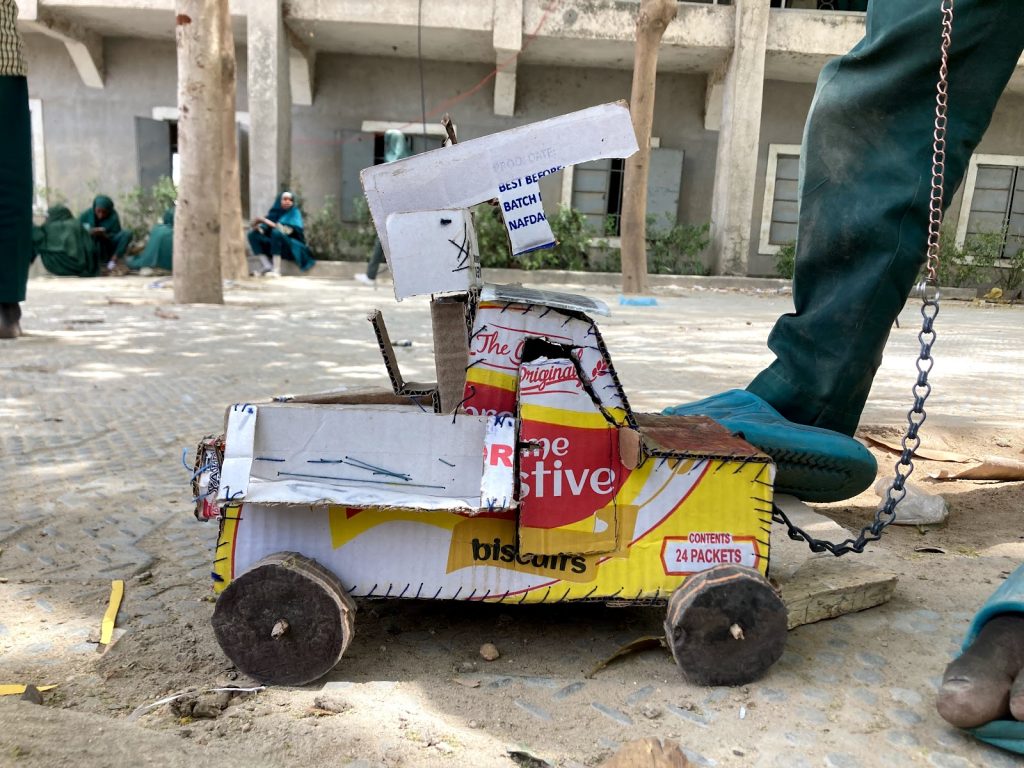 Toy truck made from reused packaging and wood, placed outdoors on sandy pavement. A person's foot in a blue shoe is visible nearby.