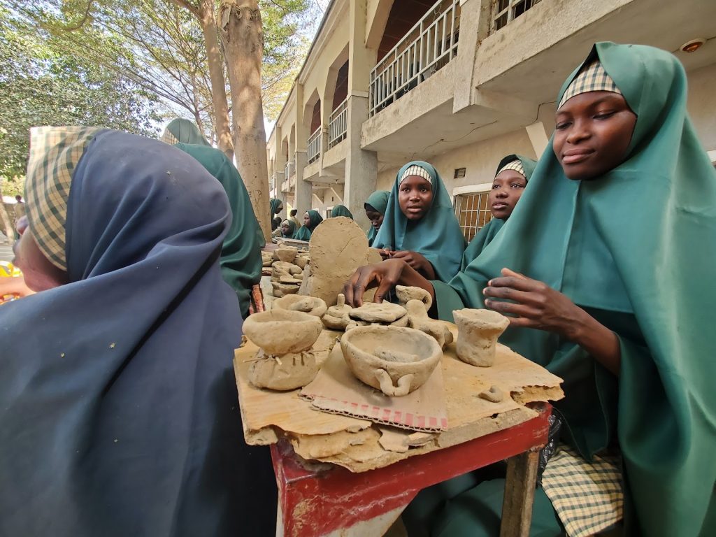 Students in green hijabs work on pottery projects outdoors, with clay pieces on a table, under a tree near a building.