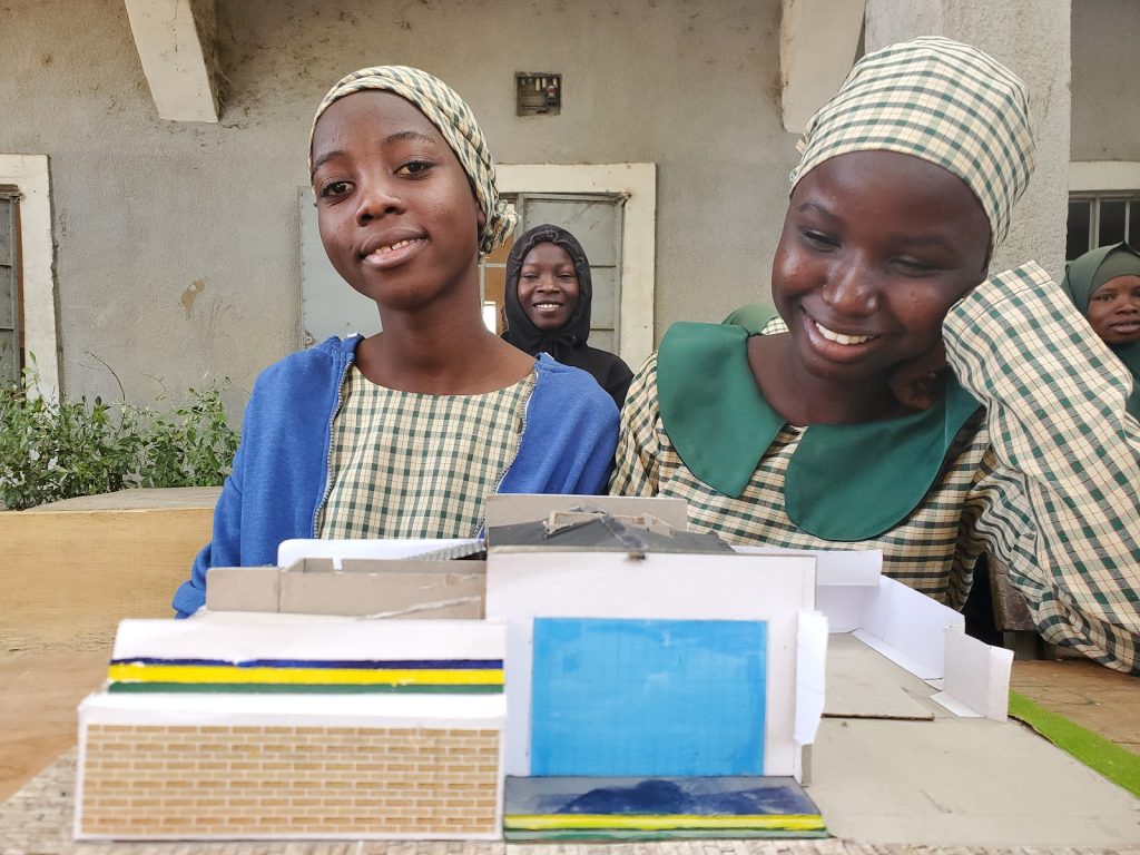 Two smiling girls in checkered uniforms present a building model at a table, while classmates look on in the background.