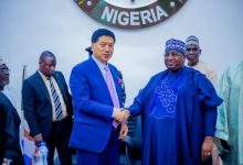 Two men in formal attire shake hands in front of a Nigerian emblem. Others are present in the background.