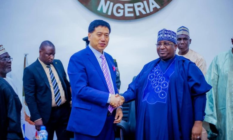 Two men in formal attire shake hands in front of a Nigerian emblem. Others are present in the background.