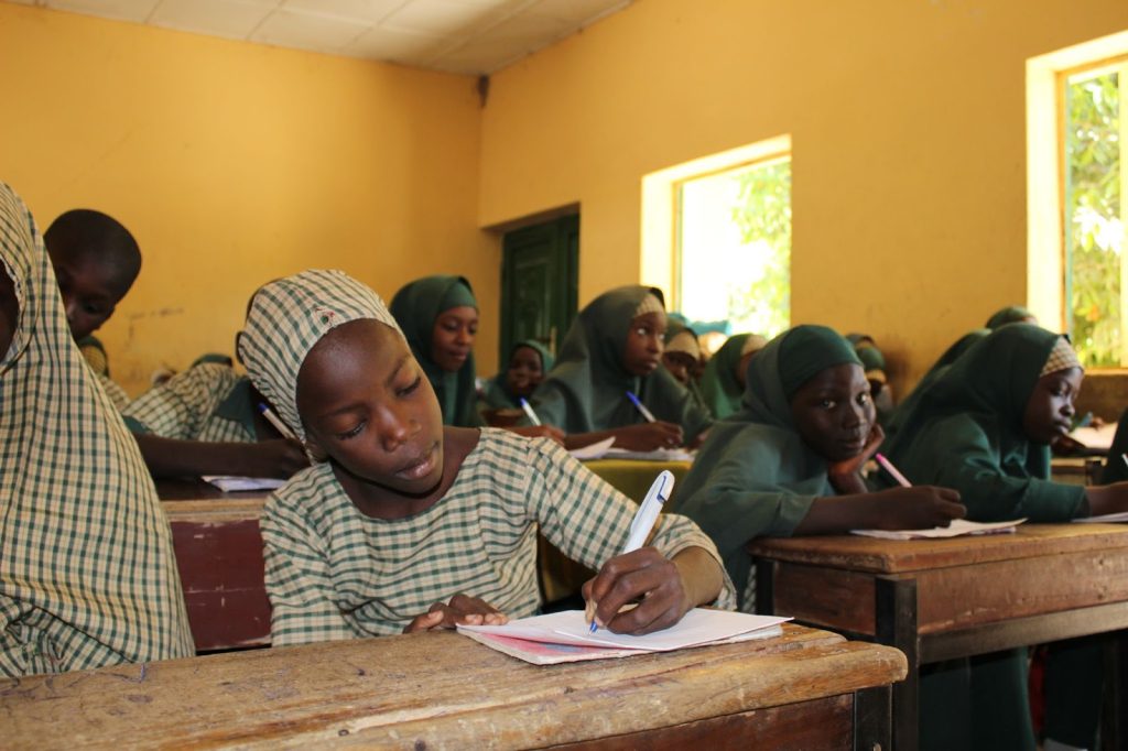 Students in a classroom, wearing green uniforms, focused on writing in notebooks with pens, near large open windows.