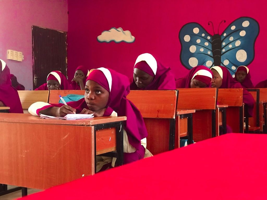 Students in pink uniforms write at wooden desks in a classroom with bright pink walls and a large butterfly mural.