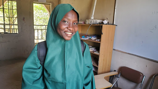 Smiling person in a green hijab and backpack stands in a classroom with shelves and a whiteboard in the background.