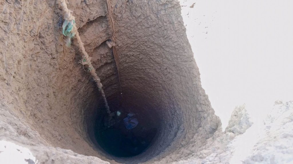 A deep, dry well with a rope hanging down, surrounded by rough, clay-like walls, viewed from above.