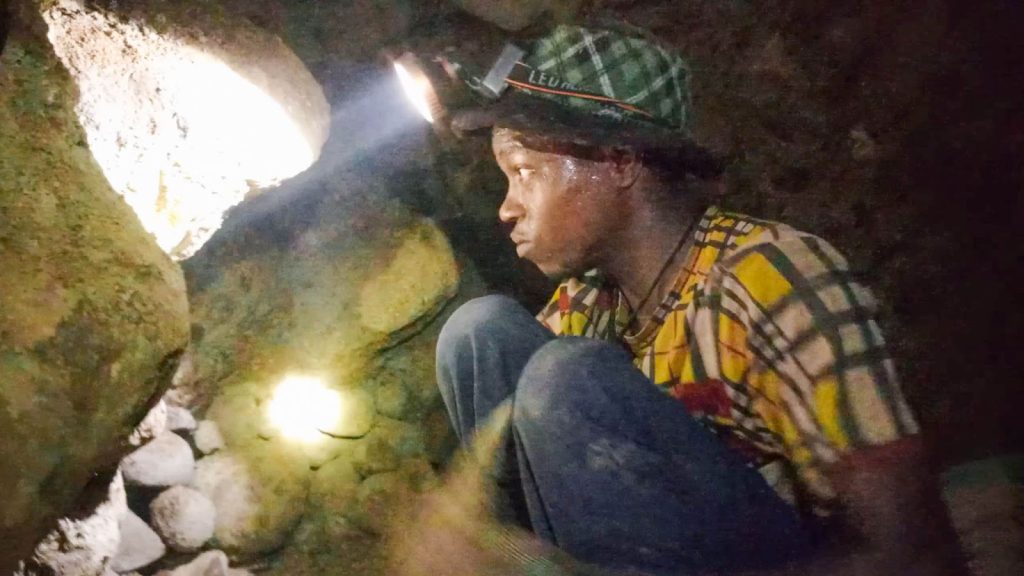 A person in a checkered shirt and headlamp sits inside a dimly lit cave, examining rocks.