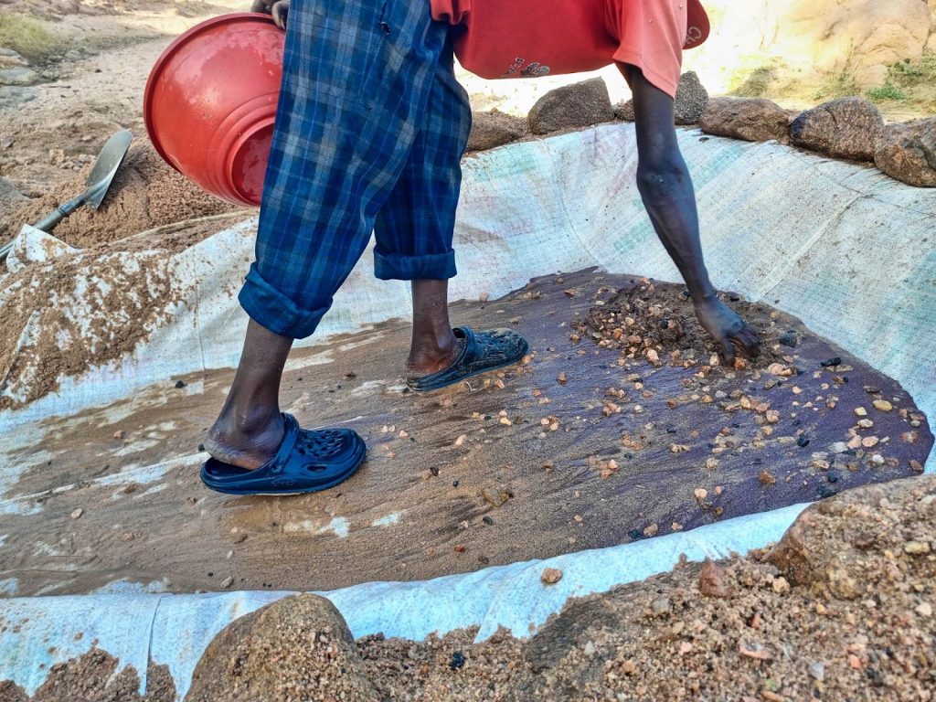 Person in casual attire processes rocks on a tarp, using a shovel and wearing sandals, with a red bucket nearby.