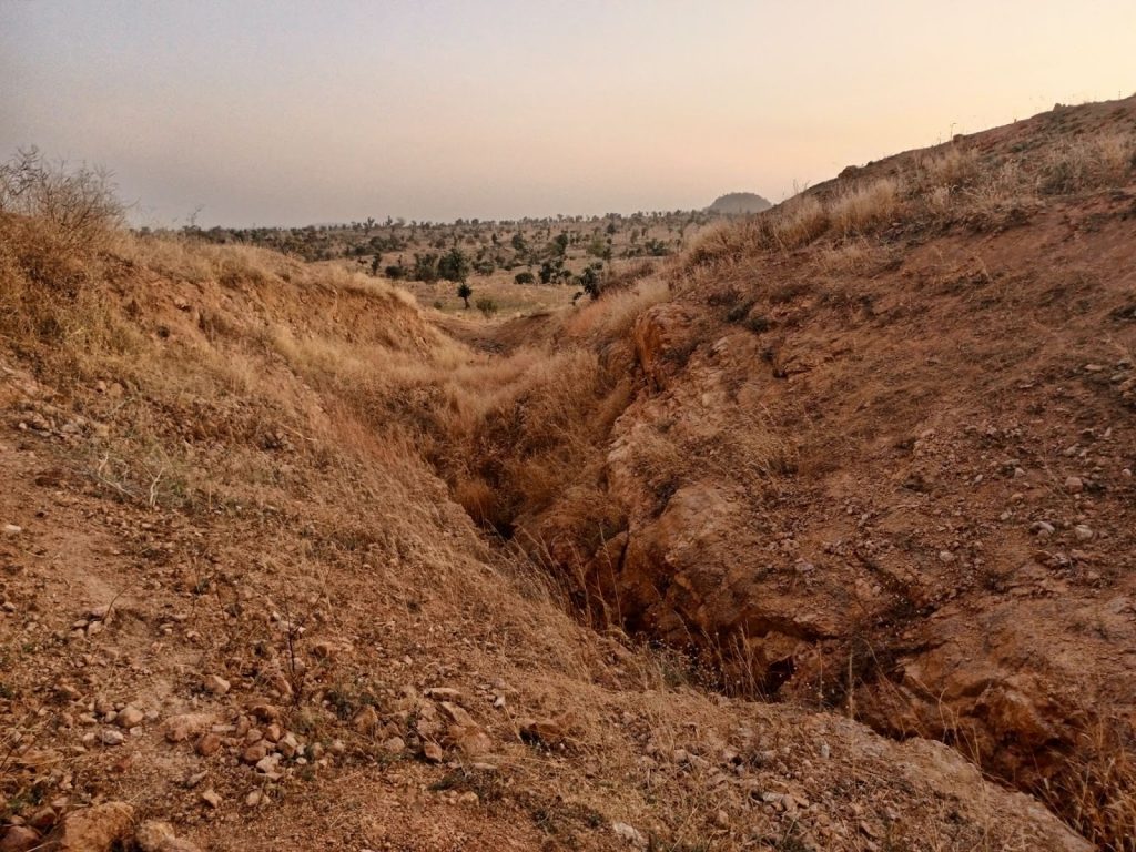 Dry terrain with a deep erosion gully surrounded by sparse grass and distant hills under a hazy sky.