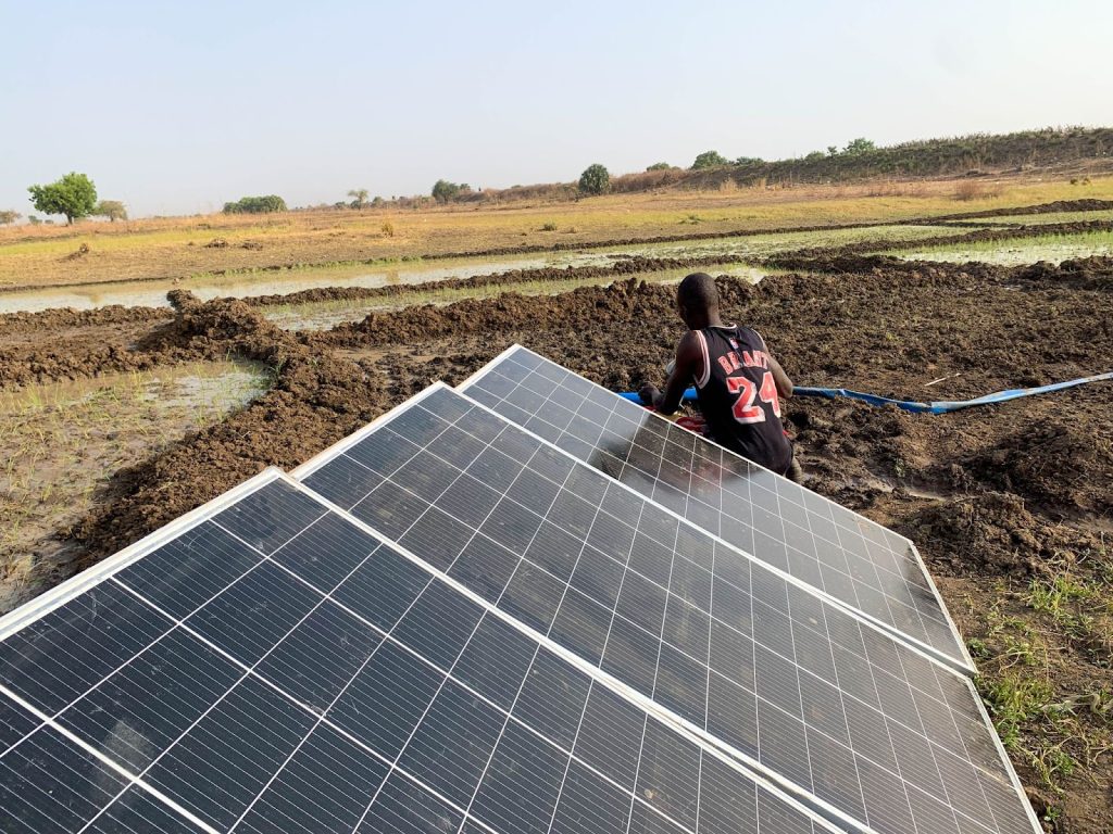 A person works next to solar panels in a muddy field, with distant trees and clear skies in the background.