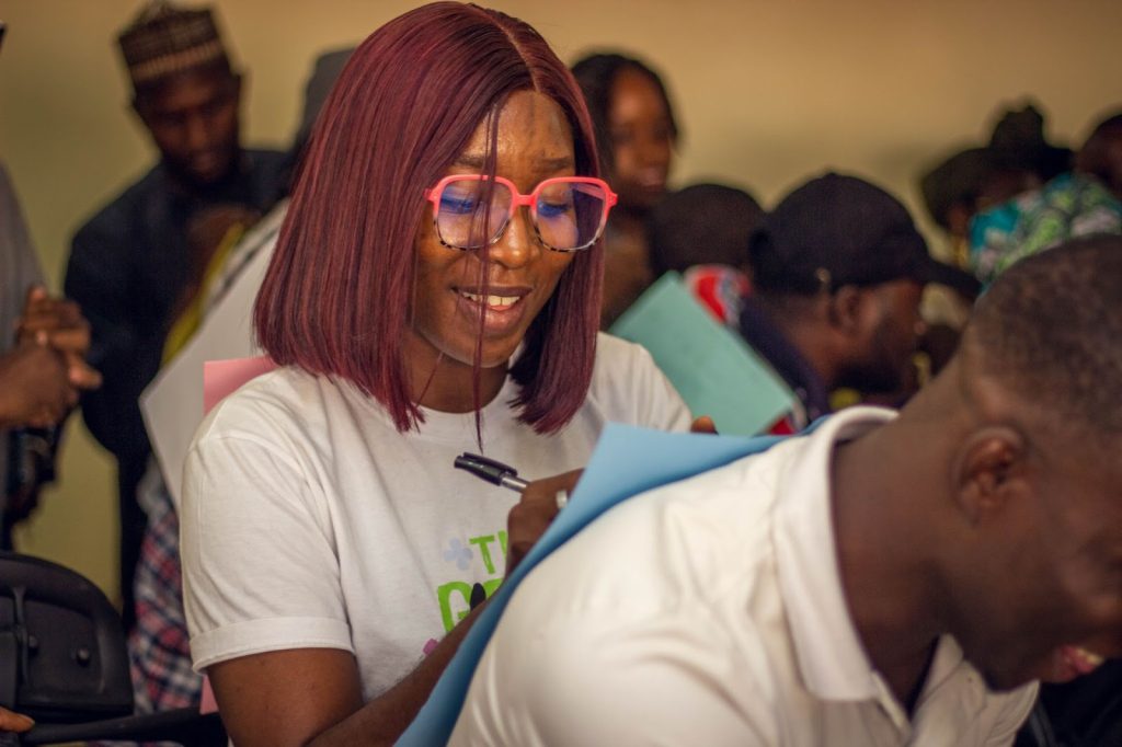 A woman with glasses smiles while writing on a blue paper in a crowded room.