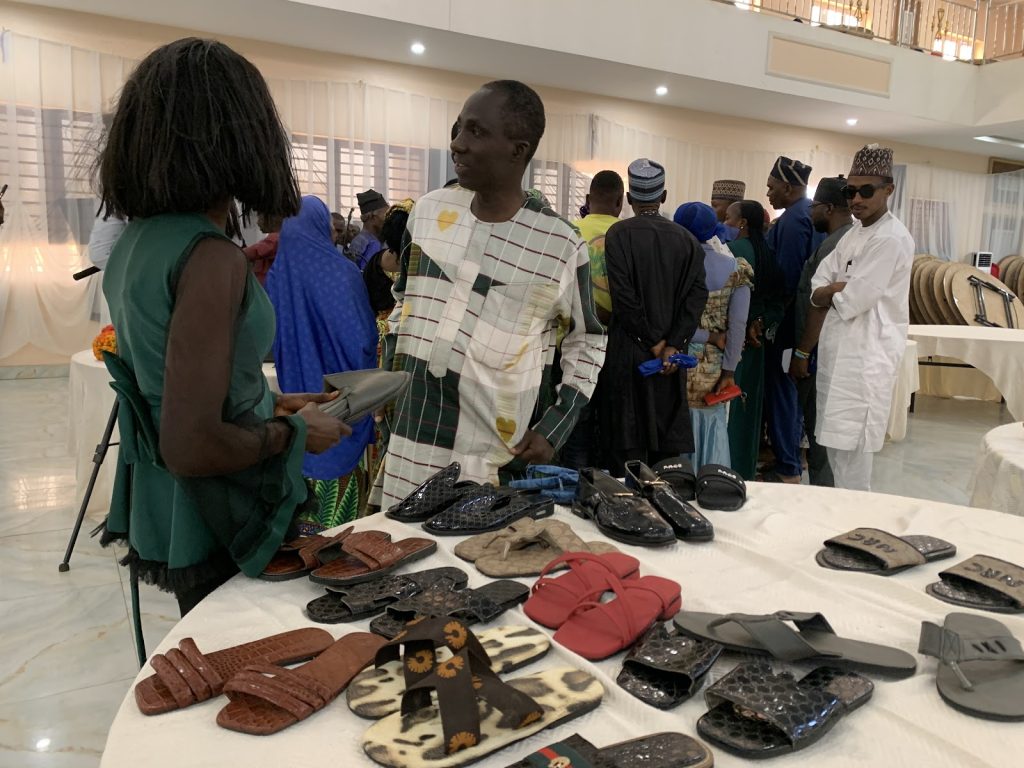 People browsing sandals and shoes on display at a market event indoors, with various footwear styles on a table in the foreground.