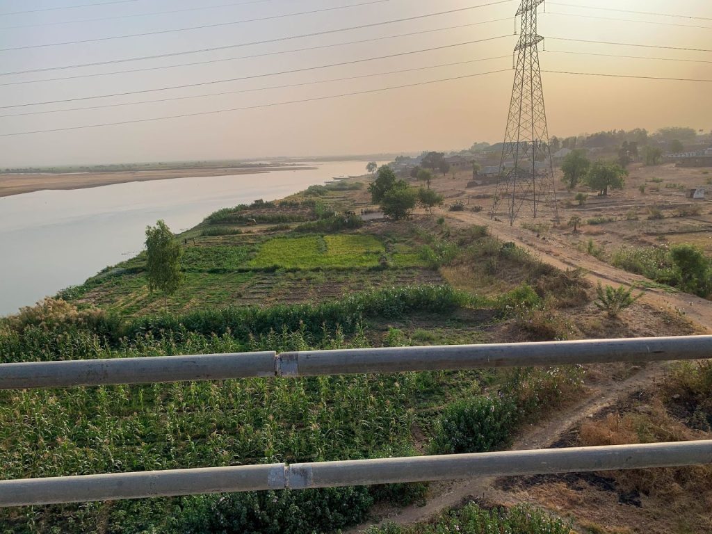 Lush green fields by a serene river under a hazy sky, with power lines and small rural houses in the background.