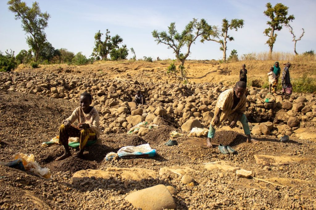 Children collecting gravel in a rocky landscape, with a few people in the background and sparse trees in a dry area.